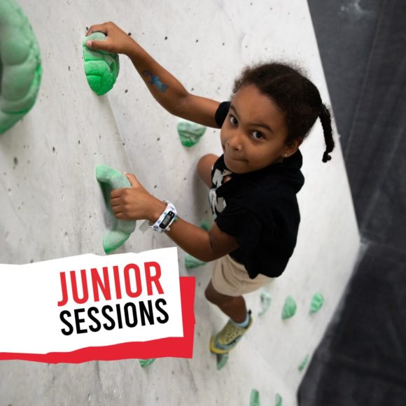 a young black female climber climbs a green route and looks into the camera during one of our junior sessions climbing classes