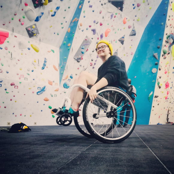 Lucy Keyworth looks at the camera from her wheelchair in front of a roped climbing wall