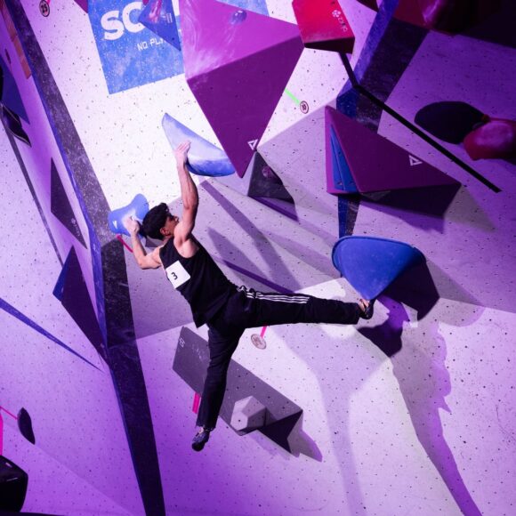Dayan Akhtar toe hooks a volume while holding a wide grip with arms above his head on a steep wall in the finals of the BMC British Bouldering Championships hosted by Depot Climbing Manchester.